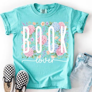 May include: A turquoise t-shirt with the words "BOOK lover" in white, surrounded by a floral design of pink, blue, and peach flowers. Black and white checkered slip-on shoes and denim shorts are next to the shirt.