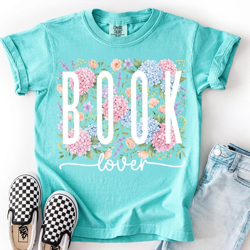 May include: A turquoise t-shirt with the words "BOOK lover" in white, surrounded by a floral design of pink, blue, and peach flowers. Black and white checkered slip-on shoes and denim shorts are next to the shirt.