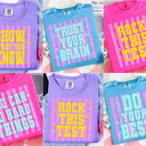 May include: A collection of colorful t-shirts in shades of purple, blue, and pink. Each shirt features a bold, retro-style graphic with motivational phrases like "Show What You Know" and "Rock This Test".