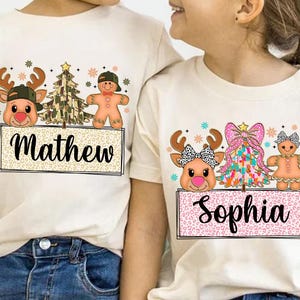 May include: Two white t-shirts with a Christmas theme. The left shirt has a camouflage Christmas tree, a gingerbread man, and a reindeer with the name "Mathew". The right shirt has a pink and white Christmas tree, a gingerbread woman, and a reindeer with the name "Sophia".
