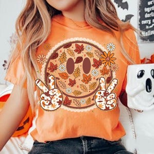 May include: Orange t-shirt featuring a smiley face design with fall leaves, flowers, and peace signs. The smiley face has brown eyes and a brown mouth. The peace signs and the smiley face are decorated with fall leaves.