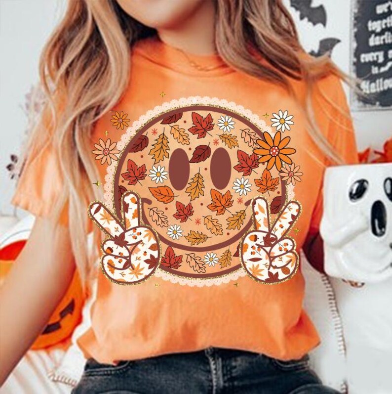 May include: Orange t-shirt featuring a smiley face design with fall leaves, flowers, and peace signs. The smiley face has brown eyes and a brown mouth. The peace signs and the smiley face are decorated with fall leaves.