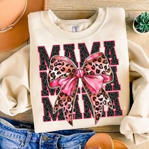 Pode incluir: Uma camisa de moletom cor creme com a palavra "MAMA" impressa em preto e um la&ccedil;o de leopardo rosa e marrom no centro.