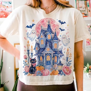 Puede incluir: Una camiseta de color crema con una ilustraci&oacute;n gr&aacute;fica de una casa encantada azul bajo una gran luna rosa. El dise&ntilde;o incluye tres fantasmas blancos, un gato negro con un lazo rosa, calabazas naranjas, flores rosas y murci&eacute;lagos volando.