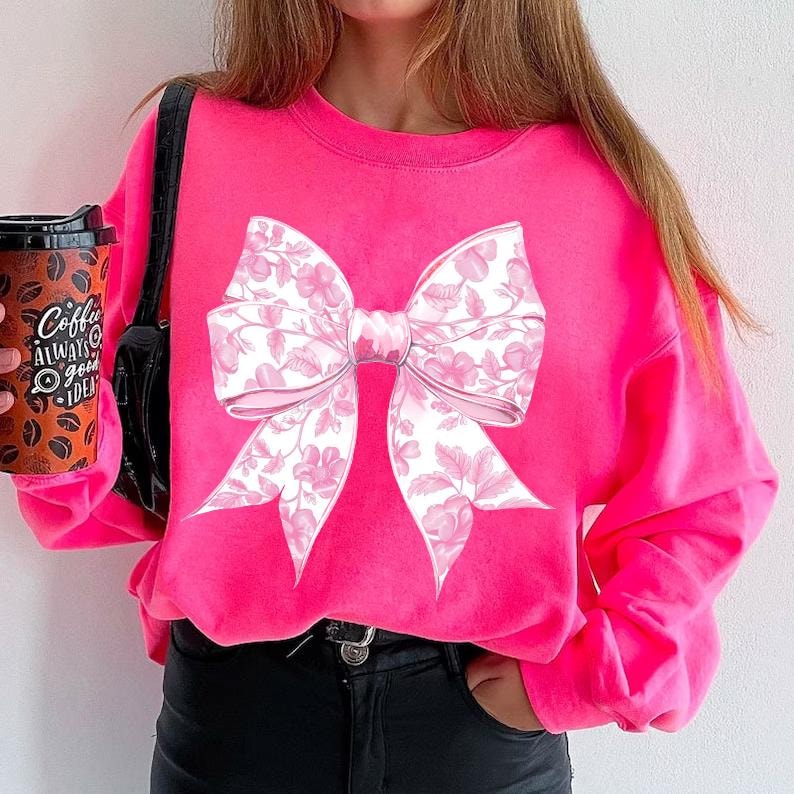 May include: A bright pink sweatshirt featuring a large white and pink bow print. The bow has a floral pattern.
