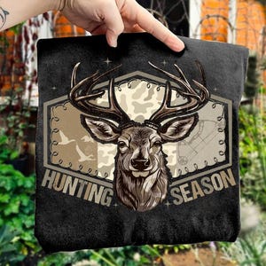 May include: Black graphic tee featuring a detailed illustration of a deer head with large antlers. The design includes a camouflage background and the words "HUNTING SEASON". The tee is held up by a hand.