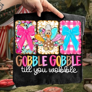 Puede incluir: Camiseta negra con un diseño colorido de Acción de Gracias. El diseño presenta tres cuadrados decorativos con un lazo rosa, un pavo y un lazo azul. El texto "GOBBLE GOBBLE till you wobble" está impreso debajo.