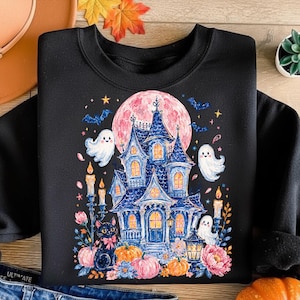 Puede incluir: Una sudadera negra de cuello redondo con un estampado gr&aacute;fico de una casa encantada azul bajo una gran luna rosa. El dise&ntilde;o incluye fantasmas blancos, murci&eacute;lagos, un gato negro con un lazo rosa, velas encendidas, calabazas naranjas y flores rosas.