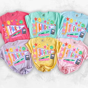 May include: A collection of five short-sleeve t-shirts in pink, mint green, light blue, lavender, and yellow. Each shirt features a graphic print of a pennant flag with school grade text, including Pre-K, Kinder, 1st Grade, 2nd Grade, 3rd Grade, and 4th Grade.