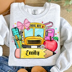 Pu&ograve; includere: Una felpa grigia con un design grafico colorato che presenta un autobus scolastico, una matita, un righello, un fiocco, una mela e il nome "Emily".