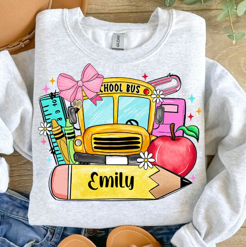 Pu&ograve; includere: Una felpa grigia con un design grafico colorato che presenta un autobus scolastico, una matita, un righello, un fiocco, una mela e il nome "Emily".