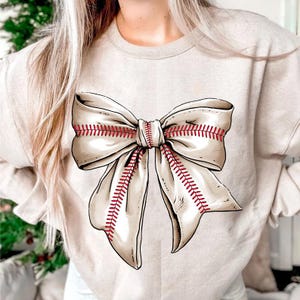 May include: A beige sweatshirt with a large white bow printed on the front. The bow has red stitching that resembles baseball stitching.