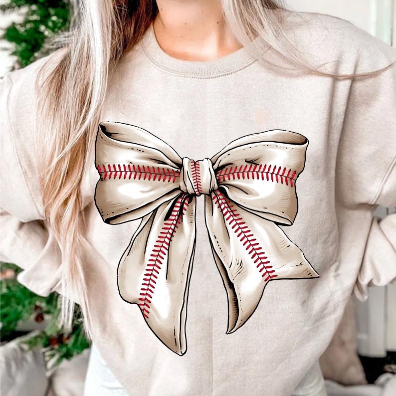 May include: A beige sweatshirt with a large white bow printed on the front. The bow has red stitching that resembles baseball stitching.
