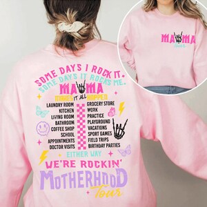 Puede incluir: Sudadera rosa con un dise&ntilde;o gr&aacute;fico que dice "Some days I rock it. Some days it rocks me. Mama makes it all happen." El gr&aacute;fico tambi&eacute;n incluye mariposas, rayos y un patr&oacute;n a cuadros. El texto "We're rockin' Motherhood Tour" tambi&eacute;n est&aacute; incluido en el dise&ntilde;o.