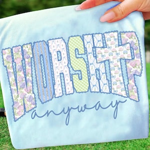 May include: A light blue t-shirt with the words "WORSHIP anyway" in a decorative font. The letters are patterned with floral, striped, and gingham designs, outlined in blue lace.