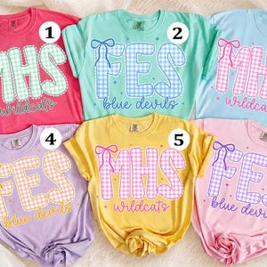 May include: Six short-sleeved t-shirts in various colors, each featuring a large gingham-patterned letter design with a bow graphic. The shirts display text including MHS Wildcats, FES Blue Devils, and FES Blue Devils, numbered one through six.