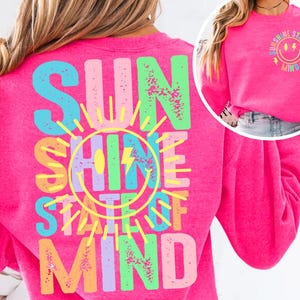 Puede incluir: Una sudadera rosa brillante con las palabras "SUNSHINE STATE OF MIND" en letras de colores en la espalda. Un diseño más pequeño en la parte delantera presenta una cara sonriente con rayos y la misma frase.