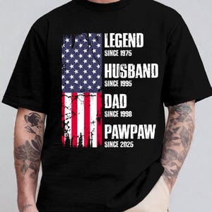K&ouml;nnte beinhalten: Schwarzes T-Shirt mit einem verblassten amerikanischen Flaggen-Design. Die Flagge ist geteilt, mit Sternen oben und Streifen unten. Text: "LEGEND SINCE 1975", "HUSBAND SINCE 1995", "DAD SINCE 1998" und "PAWPAW SINCE 2025".