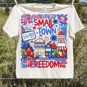 May include: Cream-colored t-shirt with a colorful graphic design. The design features the words "SMALL TOWN FREEDOM" and illustrations of buildings, fireworks, and flowers in red, white, and blue. A sign displays the number "240988".