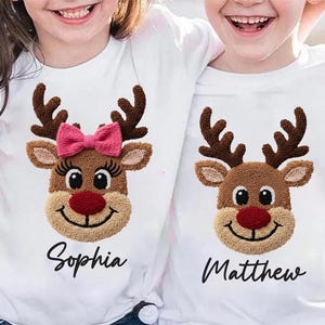 Könnte beinhalten: Zwei weiße T-Shirts mit Rentiergesichtern. Eines hat eine rosa Schleife und den Namen "Sophia" darunter. Das andere hat den Namen "Matthew" darunter. Die Rentiere haben braune Geweihe, hellbraunes Fell und rote Nasen.
