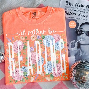 May include: Coral t-shirt with the text "i'd rather be READING" in white, framed by a floral arrangement of pink, blue, and purple flowers. The shirt is a Comfort Colors brand. A newspaper and disco ball are in the background.