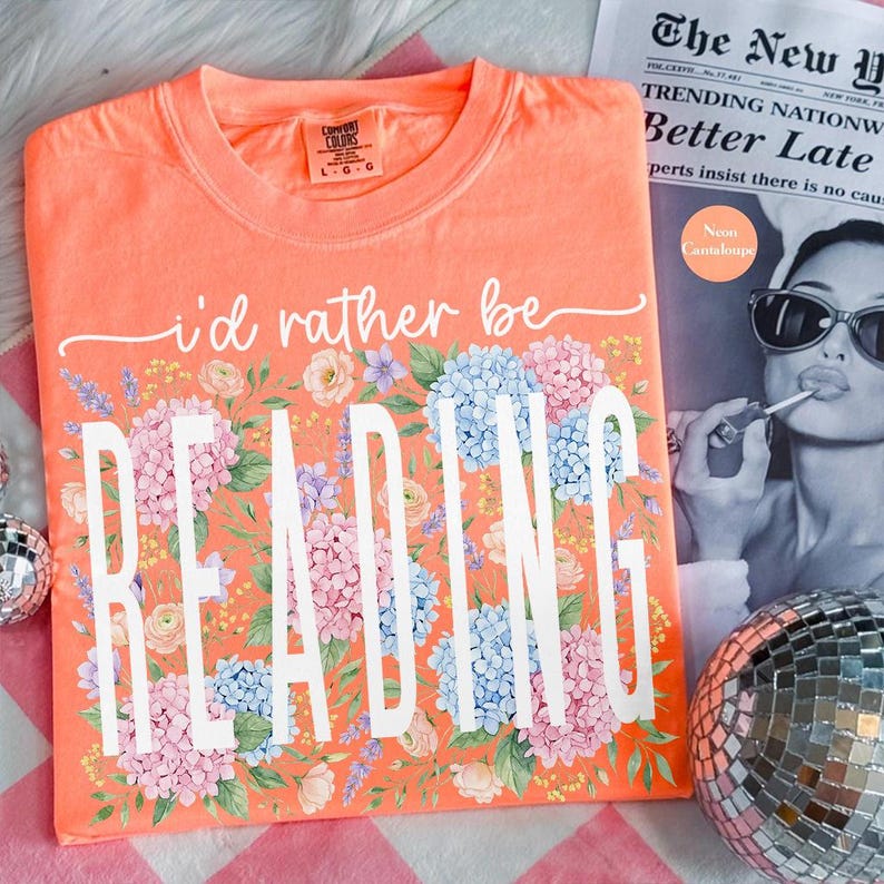 May include: Coral t-shirt with the text "i'd rather be READING" in white, framed by a floral arrangement of pink, blue, and purple flowers. The shirt is a Comfort Colors brand. A newspaper and disco ball are in the background.