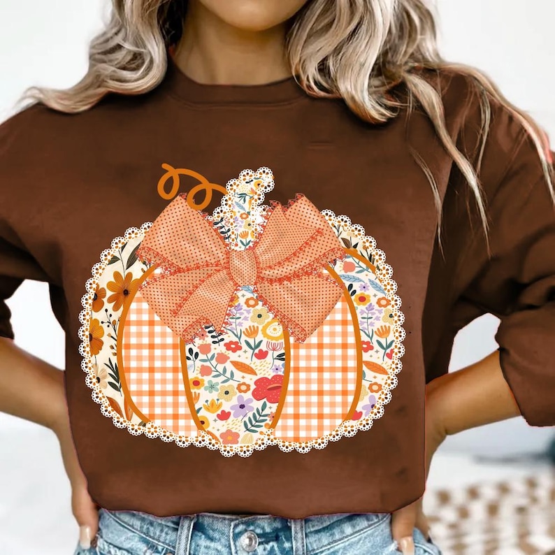 K&ouml;nnte beinhalten: Braunes Sweatshirt mit einem K&uuml;rbis-Design. Der K&uuml;rbis hat ein Blumen- und Karomuster, eine orangefarbene Schleife und einen Stiel. Das Design ist mittig auf der Vorderseite des Shirts platziert, ideal f&uuml;r den Herbst.