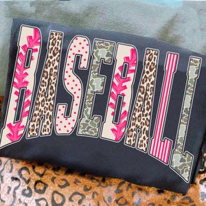 May include: A black sweatshirt with the word "BASEBALL" in a bold, multi-patterned design. The letters feature pink, leopard print, polka dots, camouflage, and striped patterns. The sweatshirt is folded on a surface with a leopard print.