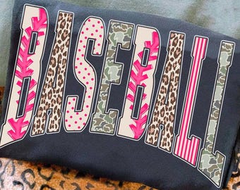May include: A black sweatshirt with the word "BASEBALL" in a bold, multi-patterned design. The letters feature pink, leopard print, polka dots, camouflage, and striped patterns. The sweatshirt is folded on a surface with a leopard print.