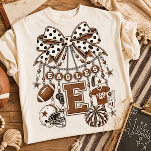 May include: A cream-colored t-shirt featuring a large graphic of a polka-dot bow hanging above a letter E, a football, a whistle, a helmet, a megaphone, a jersey labeled GAME DAY, a foam finger, and pom-poms, all connected by beaded chains.