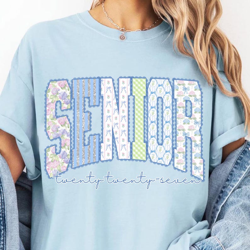May include: Light blue t-shirt with the word "SENIOR" in a decorative font. The letters are composed of floral, striped, and patterned designs. The text "twenty twenty-seven" is written below the word "SENIOR".