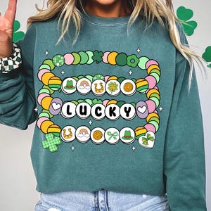 May include: Teal green sweatshirt with a graphic of a multi-colored beaded bracelet with charms. The charms include shamrocks, horseshoes, rainbows, and hats. The bracelet spells out "LUCKY" in white letters.