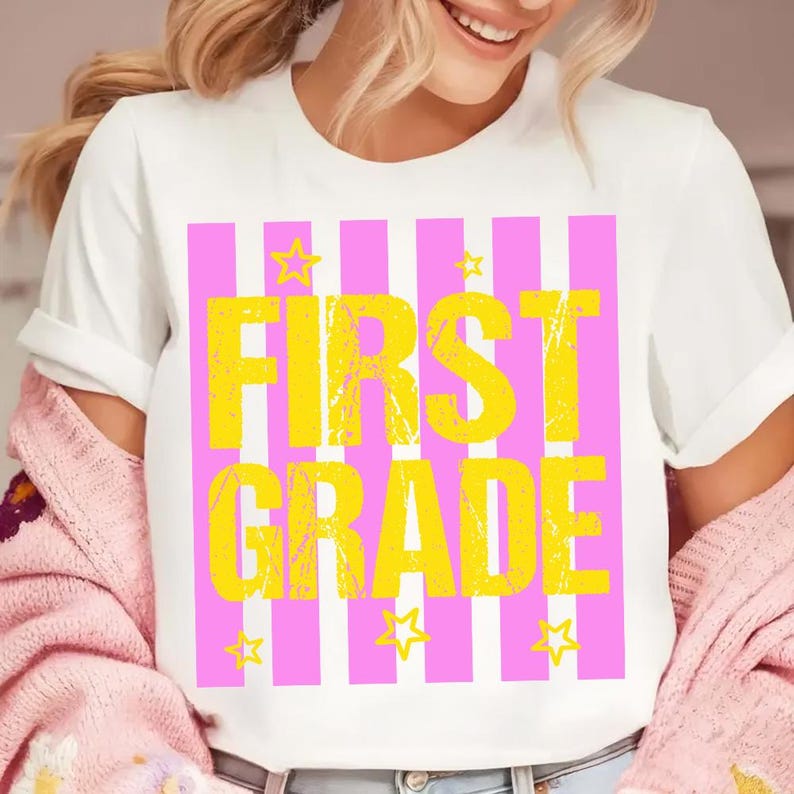 May include: White t-shirt with a pink and white striped design. The shirt features the words "FIRST GRADE" in large yellow letters, with yellow stars scattered around the text. A casual and fun back-to-school design.