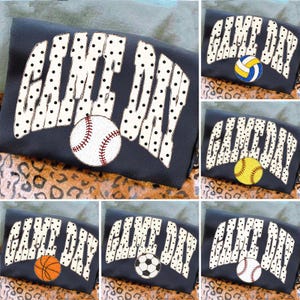 May include: A black t-shirt featuring the text GAME DAY in white letters with black polka dots. The design includes various sports balls, including a baseball, volleyball, softball, basketball, soccer ball, and another baseball, placed near the text.