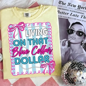 May include: A light yellow Comfort Colors t-shirt with the text "LIVING ON THAT Blue Collar DOLLAR" in a pink and blue design. The design is framed with pink bows and a white beaded border. A newspaper and disco ball are in the background.