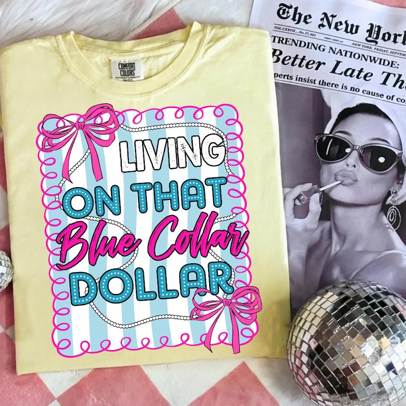 May include: A light yellow Comfort Colors t-shirt with the text "LIVING ON THAT Blue Collar DOLLAR" in a pink and blue design. The design is framed with pink bows and a white beaded border. A newspaper and disco ball are in the background.