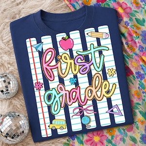 Peut inclure: T-shirt bleu marine avec un motif color&eacute; "First Grade". Le motif comprend une pomme rose, un bus scolaire jaune, un crayon et d'autres graphiques sur le th&egrave;me de l'&eacute;cole. Les mots sont dans une police multicolore ludique.