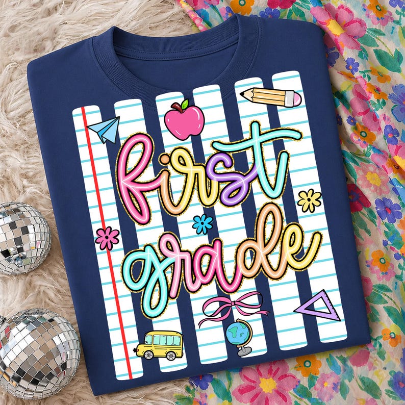 Peut inclure: T-shirt bleu marine avec un motif color&eacute; "First Grade". Le motif comprend une pomme rose, un bus scolaire jaune, un crayon et d'autres graphiques sur le th&egrave;me de l'&eacute;cole. Les mots sont dans une police multicolore ludique.