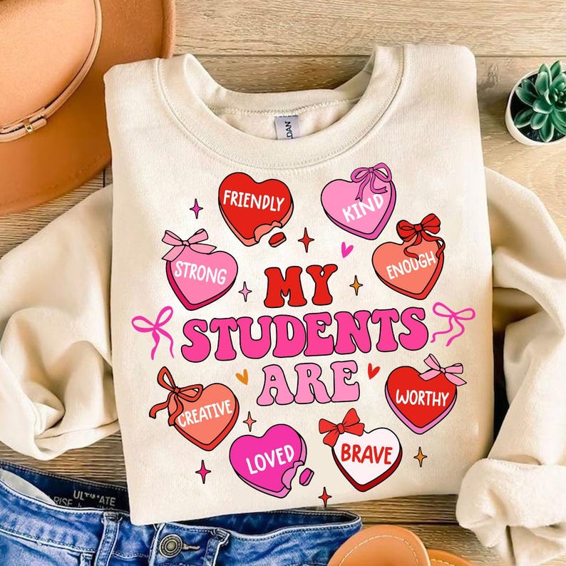 May include: A cream-colored sweatshirt with a pink and red graphic design. The graphic features hearts with words like "strong", "creative", "loved", and "brave". The text "My Students Are" is in large pink letters.