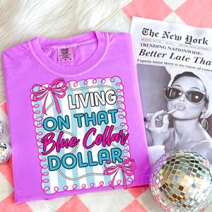 May include: A lilac Comfort Colors t-shirt with the phrase "LIVING ON THAT Blue Collar DOLLAR" in a decorative frame. The design includes pink bows and a blue and white striped background. A newspaper and disco ball are in the background.