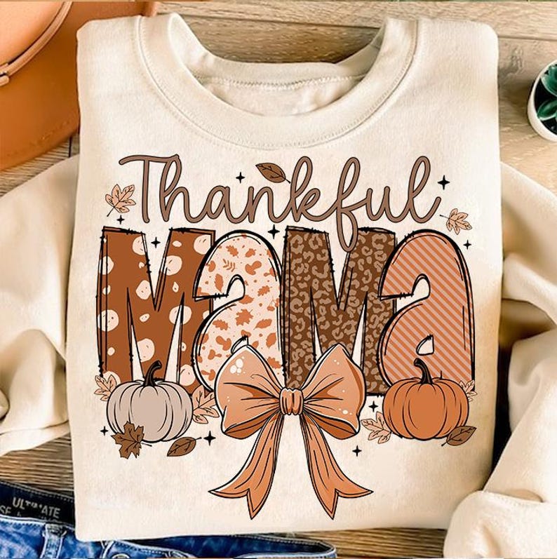 Op de afbeelding: Een beige sweatshirt met de tekst "Thankful Mama" in bruine letters met een patchwork-ontwerp. De letters zijn versierd met bruine en witte stippen, luipaardprint en strepen. Onder de tekst bevindt zich een grote bruine strik met een wit centrum. Twee kleine oranje pompoenen met groene stelen bevinden zich aan weerszijden van de strik.