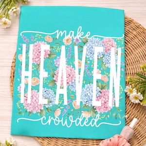May include: A turquoise decorative sign with the words "make HEAVEN crowded" in white. The sign is decorated with pink and blue hydrangea flowers and green leaves. The sign is on a woven surface.
