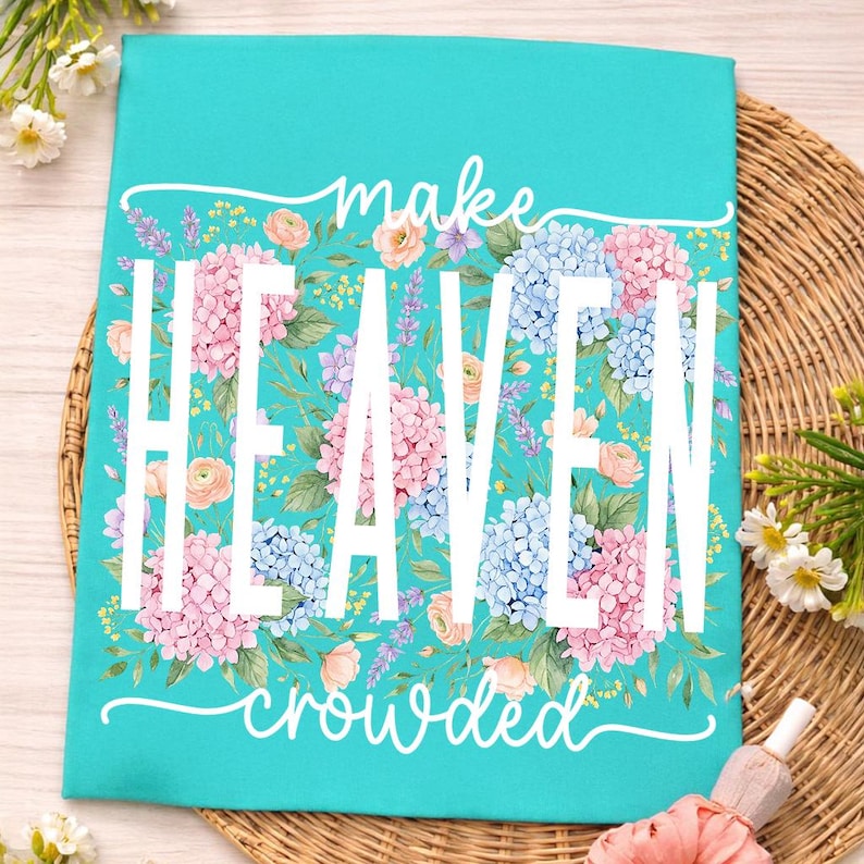 May include: A turquoise decorative sign with the words "make HEAVEN crowded" in white. The sign is decorated with pink and blue hydrangea flowers and green leaves. The sign is on a woven surface.
