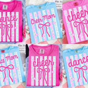 May include: A collection of six t-shirts in pink and light blue. Each shirt features a design with vertical stripes, a bow, and text. The text includes "dance", "Cheer Mom", and "cheer".