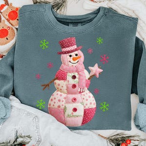 May include: A teal sweatshirt with a patchwork snowman design. The snowman wears a top hat, scarf, and holds a star wand. Pink and green snowflakes are scattered around the snowman, creating a festive winter theme.