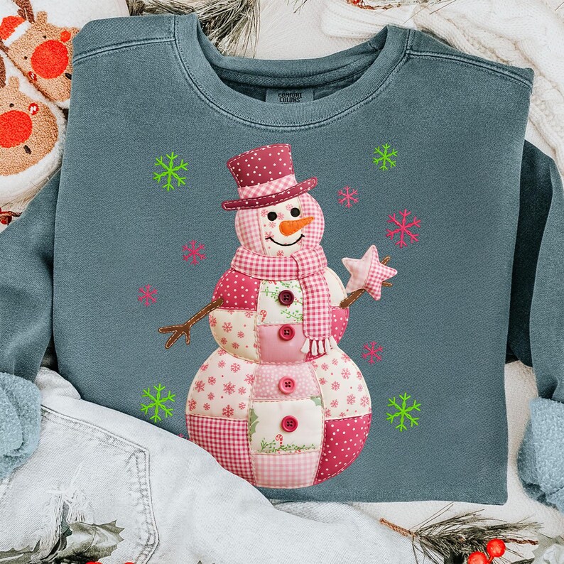 May include: A teal sweatshirt with a patchwork snowman design. The snowman wears a top hat, scarf, and holds a star wand. Pink and green snowflakes are scattered around the snowman, creating a festive winter theme.
