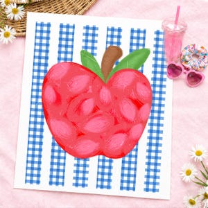 May include: A vibrant red apple illustration with green leaves and a brown stem, set against a blue and white checkered background. The artwork is on a white surface, with a pink drink, heart-shaped sunglasses, and a floral scrunchie nearby.