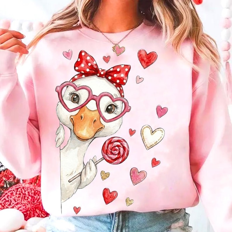 May include: Pink sweatshirt featuring a cartoon goose wearing heart-shaped glasses and a red polka dot bow. The goose holds a lollipop and is surrounded by heart graphics. A cute and whimsical design for Valentine's Day or a gift.