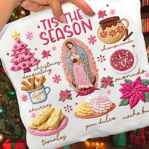 May include: White fabric with embroidered Christmas-themed designs. Features text "'Tis the Season" and illustrations of a pink Christmas tree, churros, a religious figure, and various food items. Includes the words "Christmas decorating", "churros", "tamales", and "pan dulce".