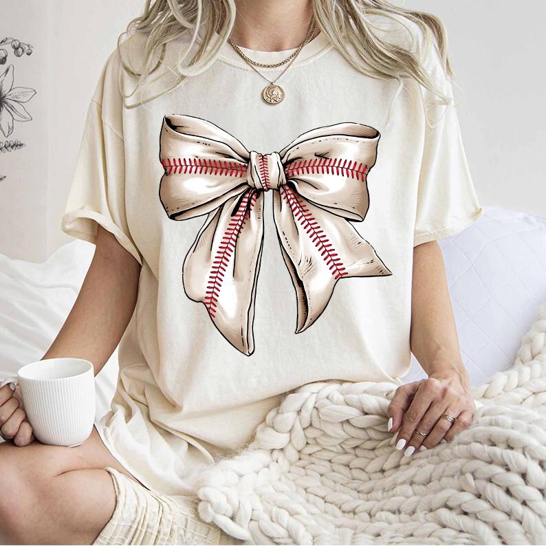 May include: A white t-shirt with a large beige bow printed on the front. The bow has red stitching that resembles a baseball stitch.